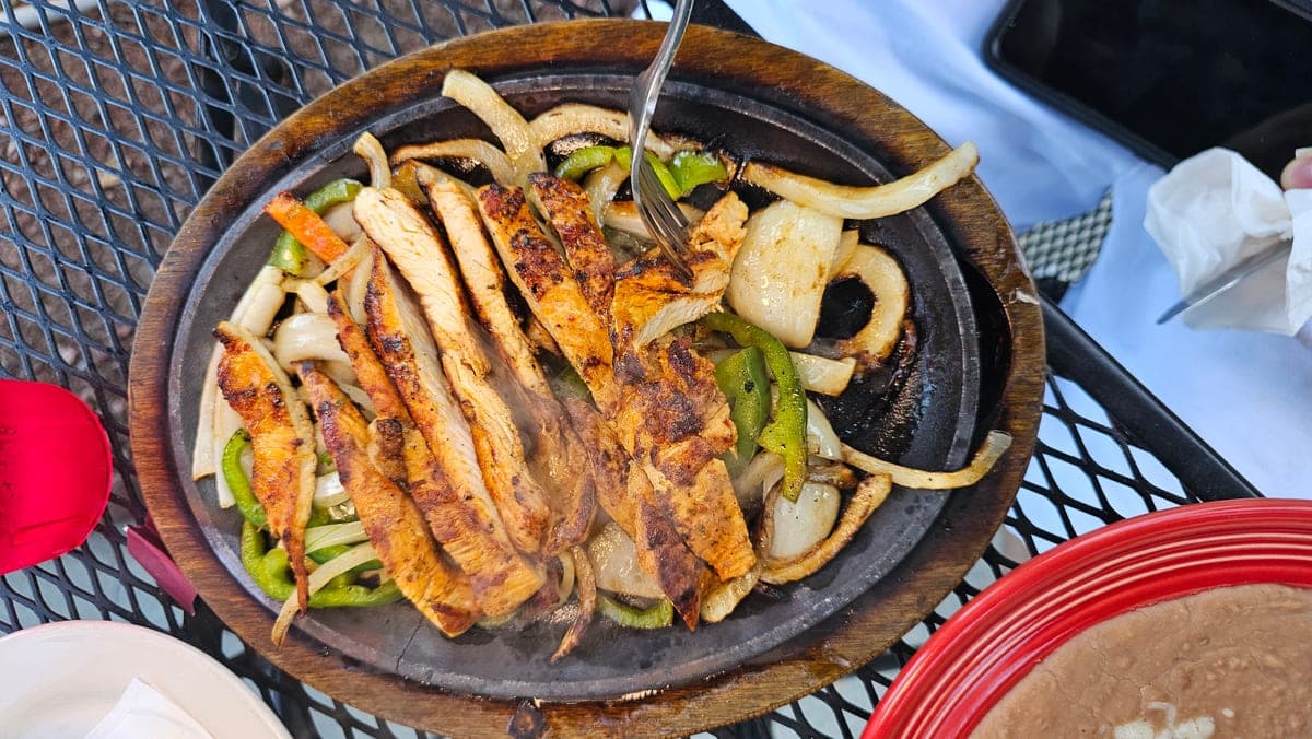 A phone photo of chicken fajitas served in a cast-iron skillet on a patio table, with a fork, napkins and other dishes around it.
