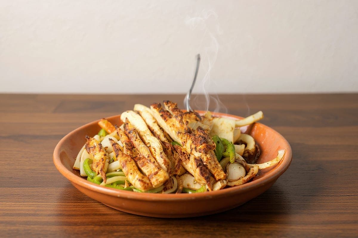 The same fajitas rendered in a shallow terracotta bowl on a polished wooden table. The food is the same; the cast-iron skillet it was served in is gone.