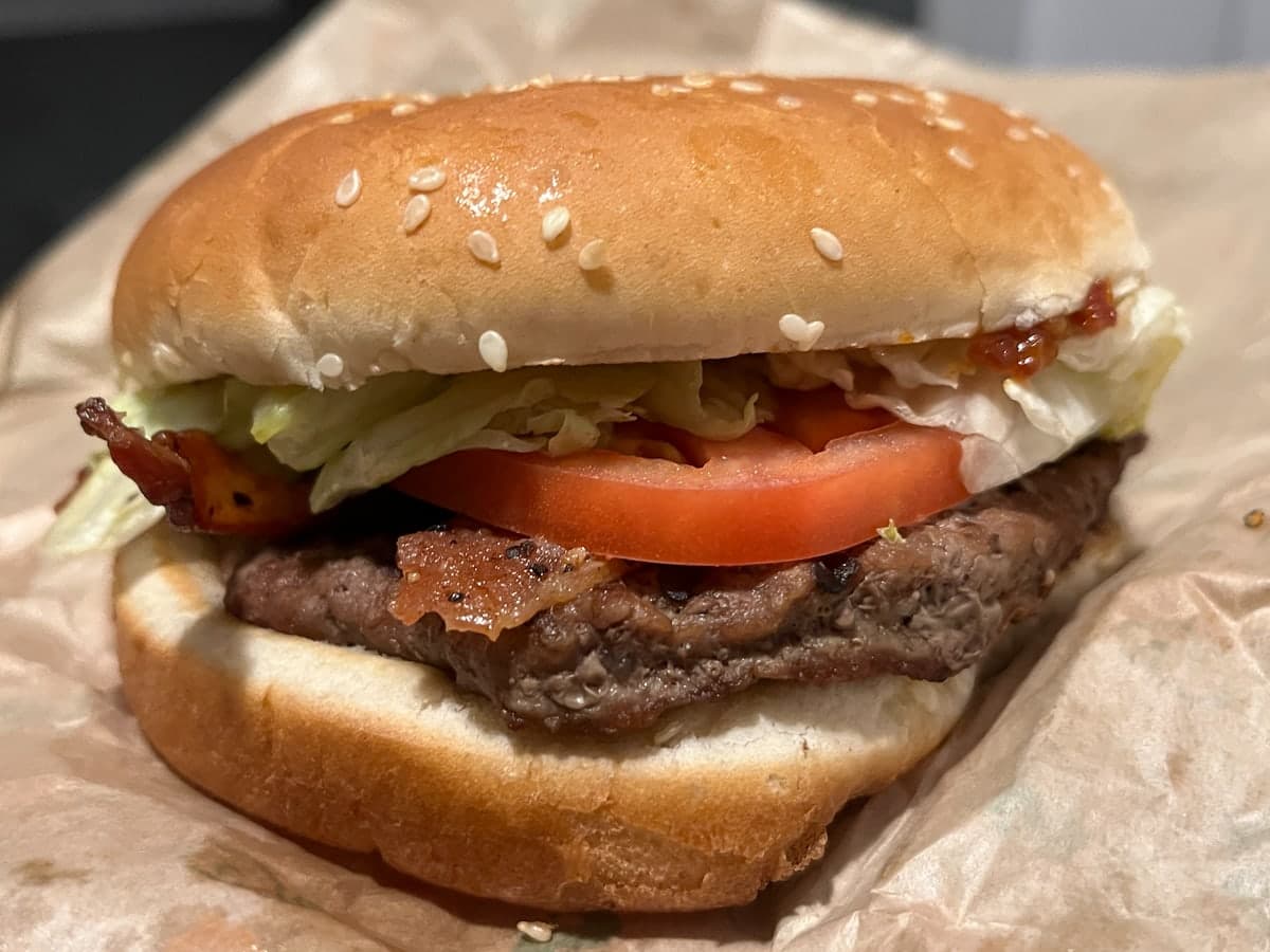 A close-up phone photo of a bacon cheeseburger on crumpled brown parchment paper under dim indoor light. The bun runs almost to the left and right edges of the frame, with no margin around it.