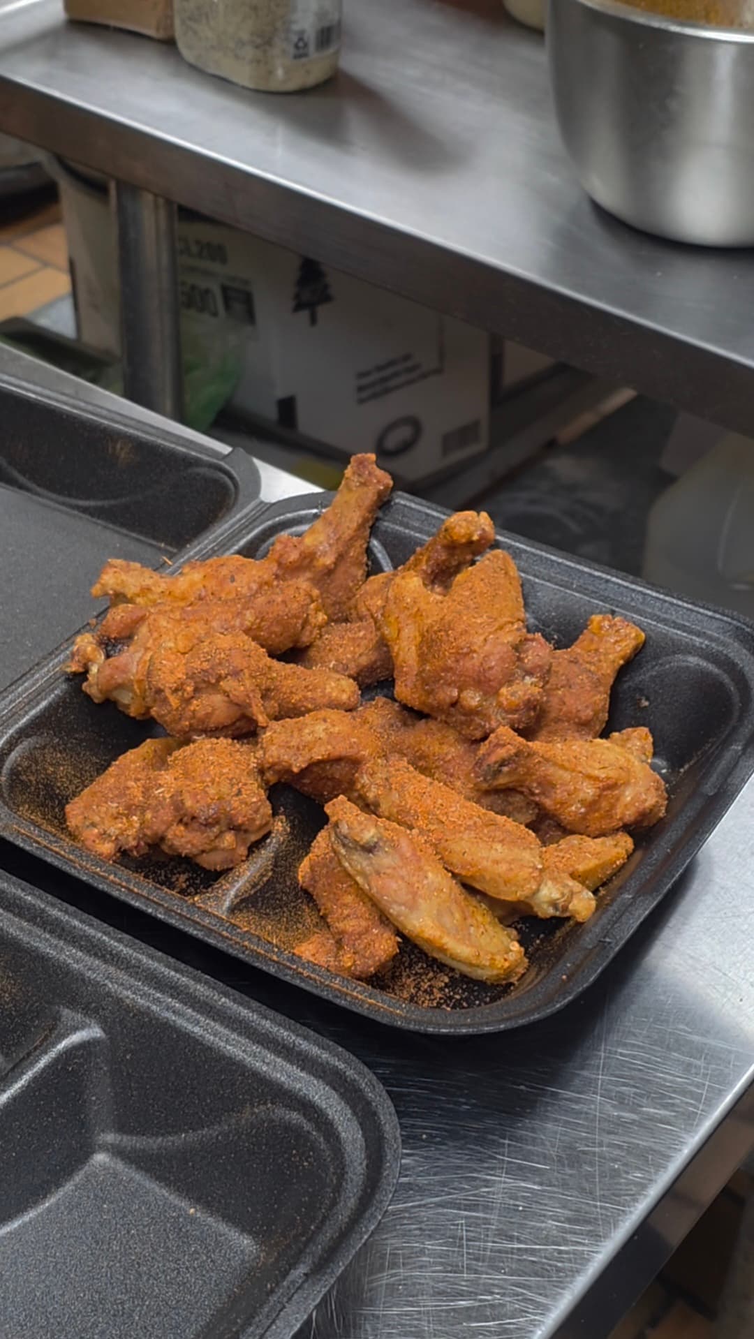 Phone photo of seasoned fried wings in a foam takeout tray on a stainless steel kitchen counter