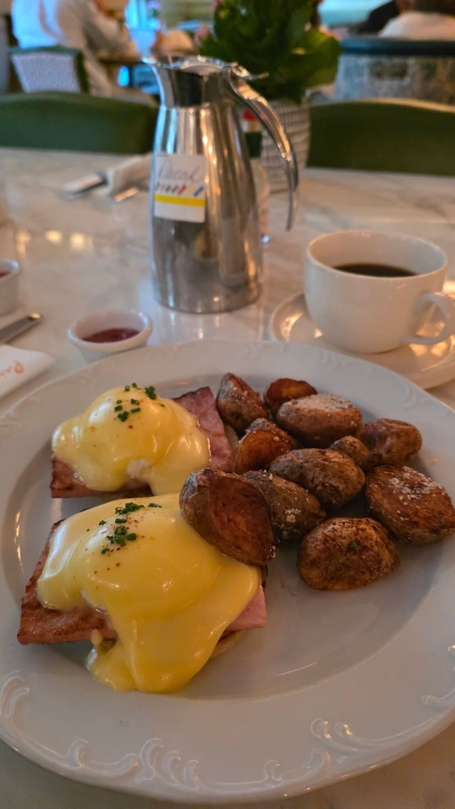 Phone photo of Eggs Benedict with roasted potatoes on a marble table, with a coffee cup and a steel carafe in the background
