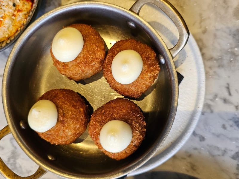 Overhead phone photo of four fried croquettes in a small steel pan, each topped with a dollop of white sauce